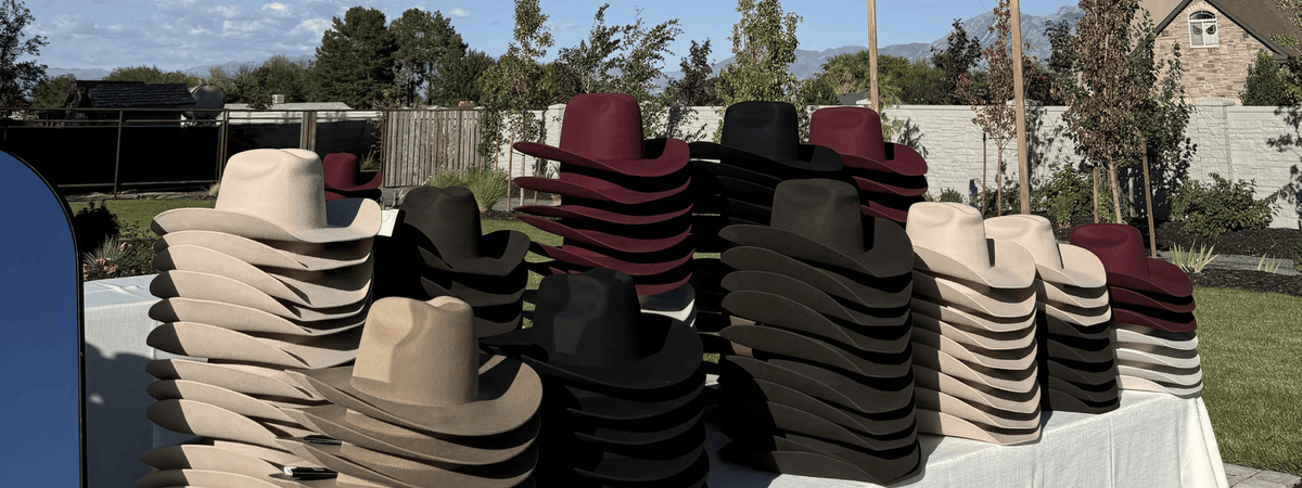 Stacks of cowboy hats in multiple colors, including beige, black, brown, and burgundy, displayed outdoors on a white-covered table in a backyard setting at a custom hat bar event.