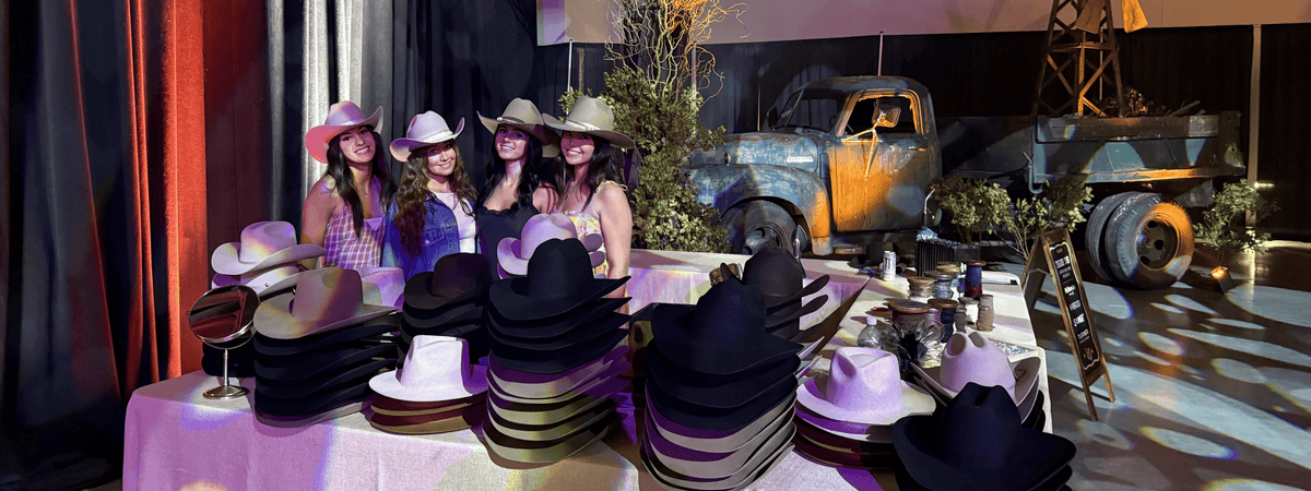 A group of women wearing cowboy hats pose behind a table covered with stacks of cowboy hats, set in a decorated event space with a vintage truck, windmill, and dramatic colored lighting at a custom hat bar event.
