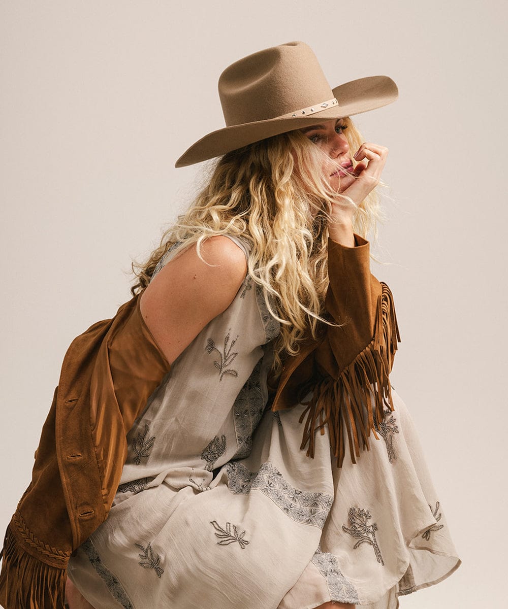 Woman wearing a brown cowboy hat and brown jacket with fringe on a plain background #color_brown