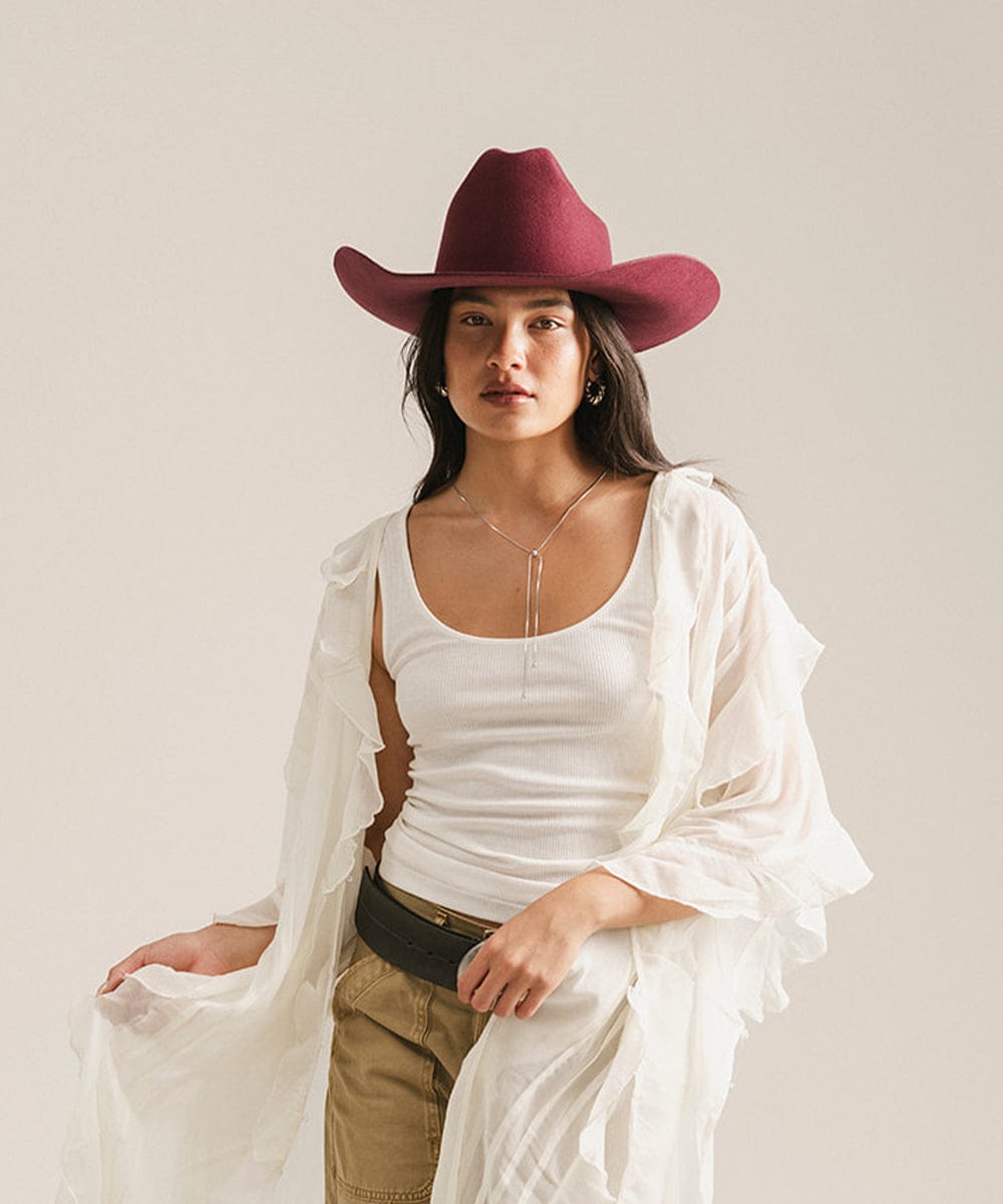 Woman wearing a white top, beige pants, and a maroon cowboy hat against a plain background #color_merlot