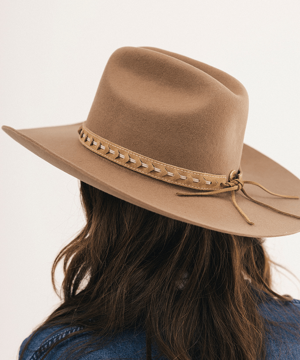 A close-up back view of a person wearing a tan wide-brim fedora hat with a matching band decorated with small metallic studs and a tie detail, against a plain light background #color_camel