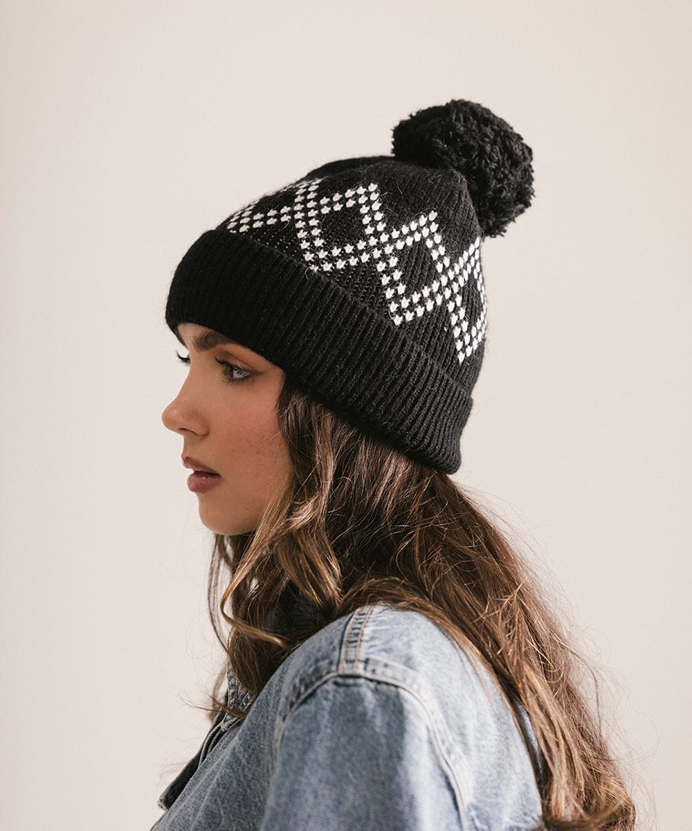 Woman wearing a black knit beanie with white geometric pattern and pom-pom on a plain background #color_black