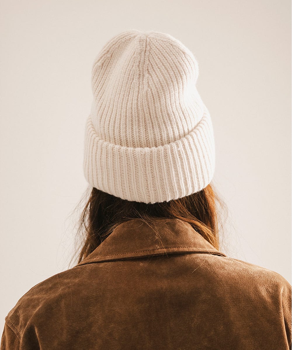 Person wearing an off white knit beanie and brown jacket, facing behind against a plain background #color_off white