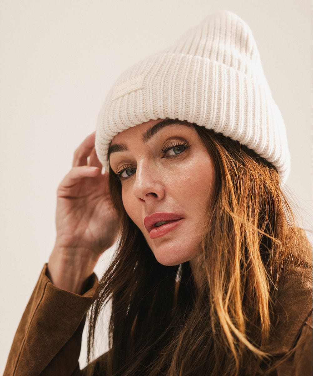 Woman wearing an off white knit beanie and brown coat against a plain background #color_off white