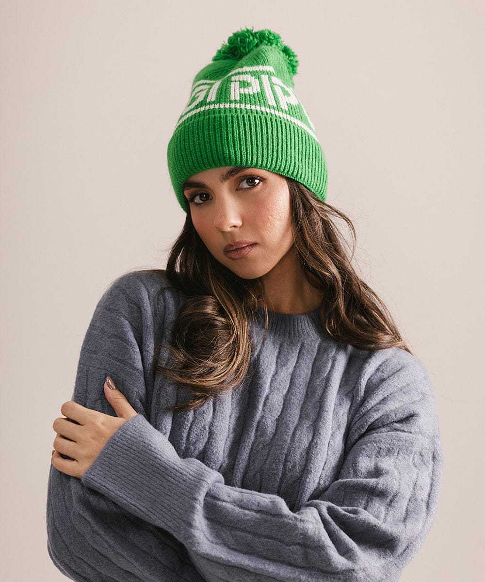 Woman wearing an evergreen colored knit beanie with pom-pom and 'GIGI PIP' branding and a blue sweater against a plain background 
