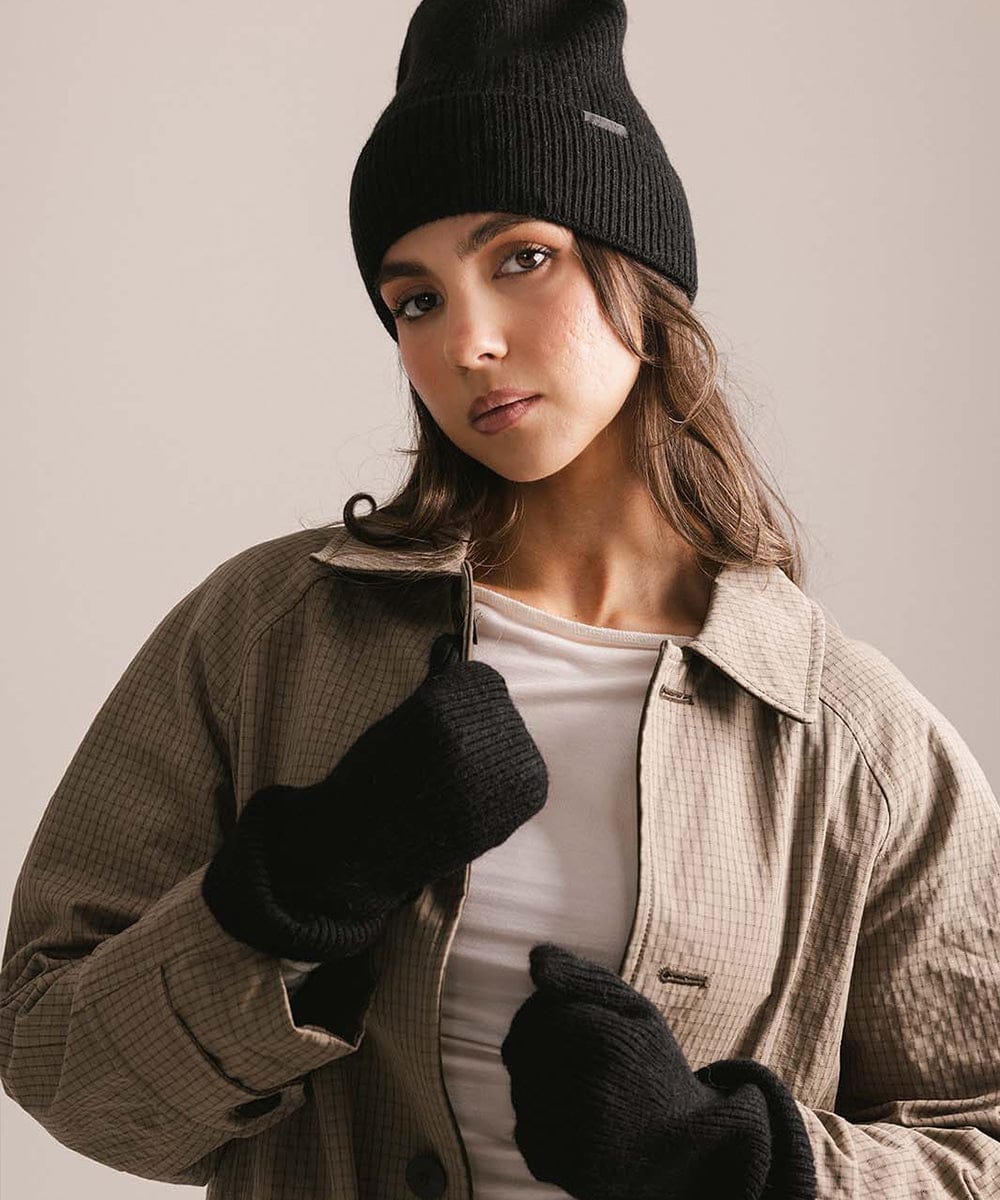 Woman wearing a black knit beanie, gloves, and a beige coat against a neutral background #color_black