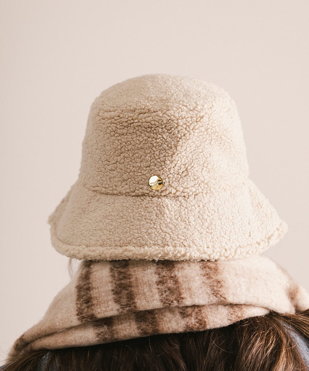 Woman wearing a cream sherpa bucket hat and a black coat against a plain background #color_cream
