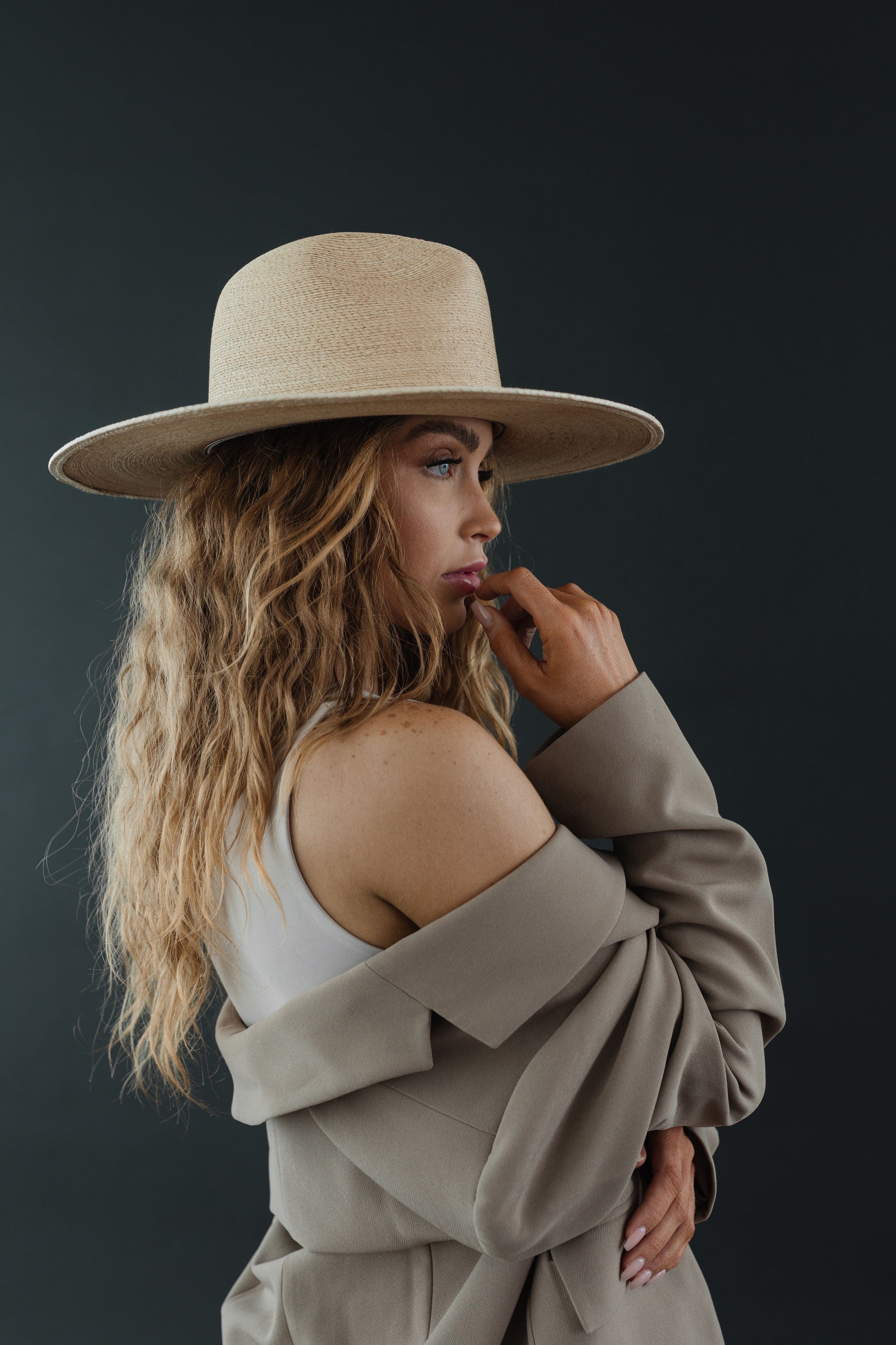 Woman wearing a toasted colored straw hat and a white top with a grey coat, facing sideways against a dark grey background #color_toasted
