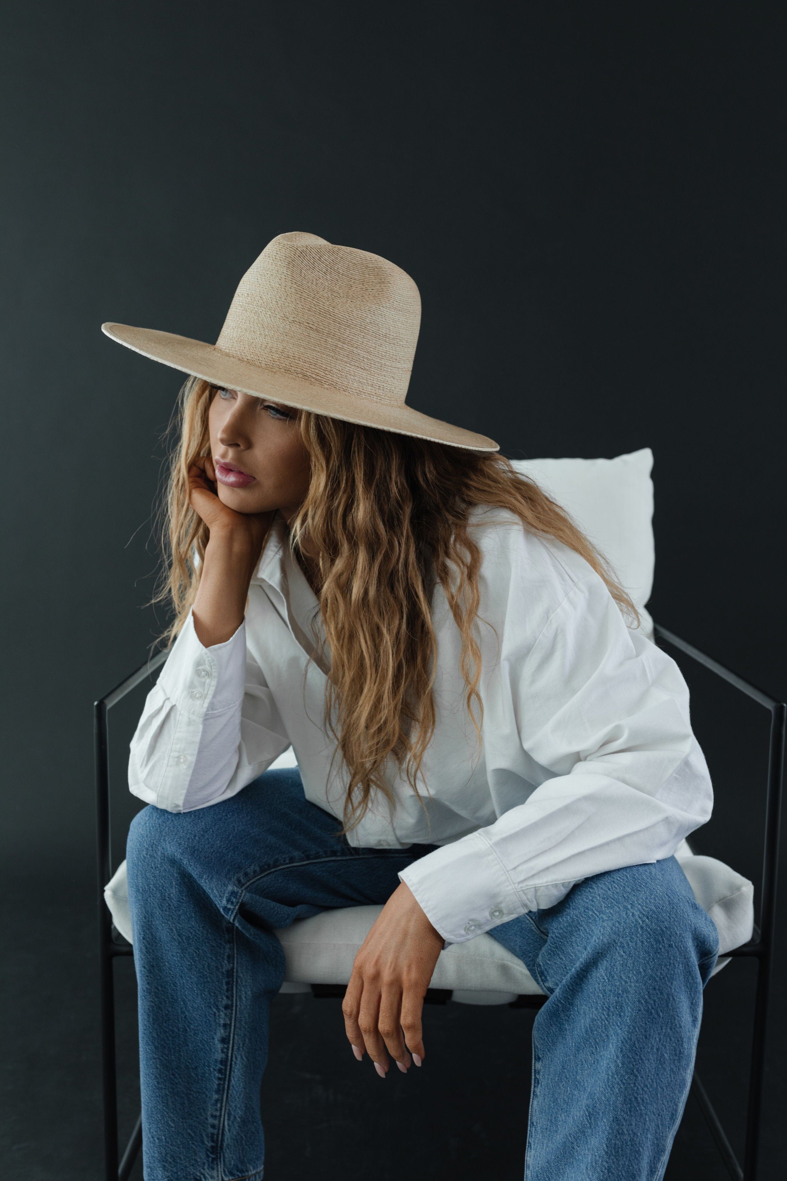 Woman wearing a toasted colored straw hat, a white shirt and blue jeans, sitting on a chair against a dark grey background #color_toasted