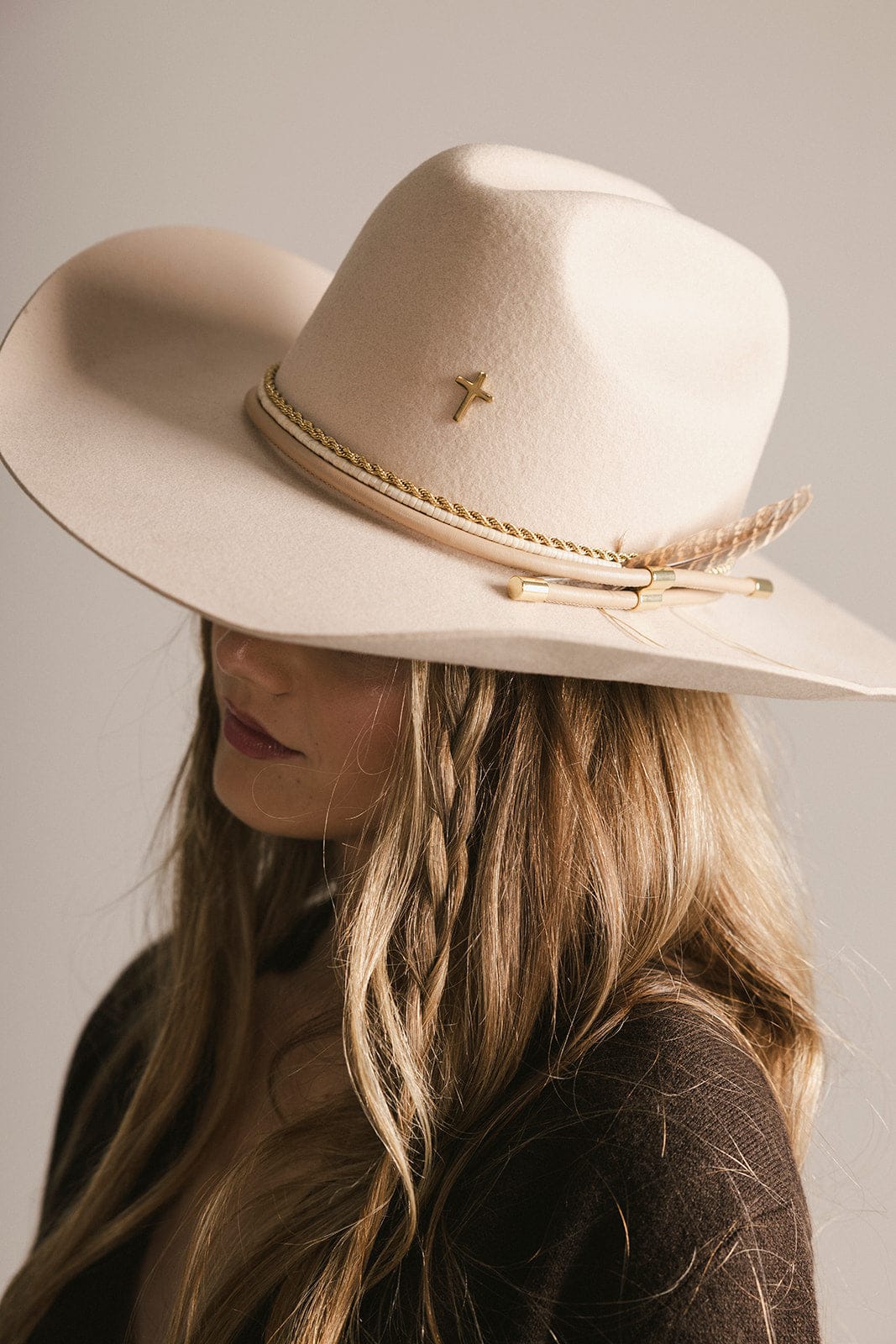 Person wearing a beige cowboy hat with a gold band and cross on a neutral background #color_gold