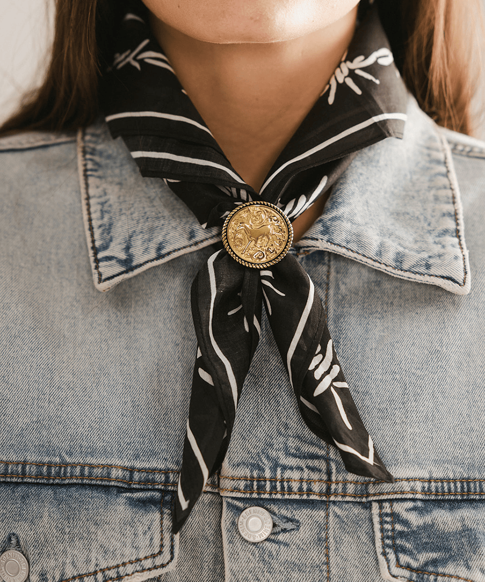 Person wearing a black and white patterned scarf with a gold coin Horse Concho Bandana Slide on a denim jacket. #color_gold