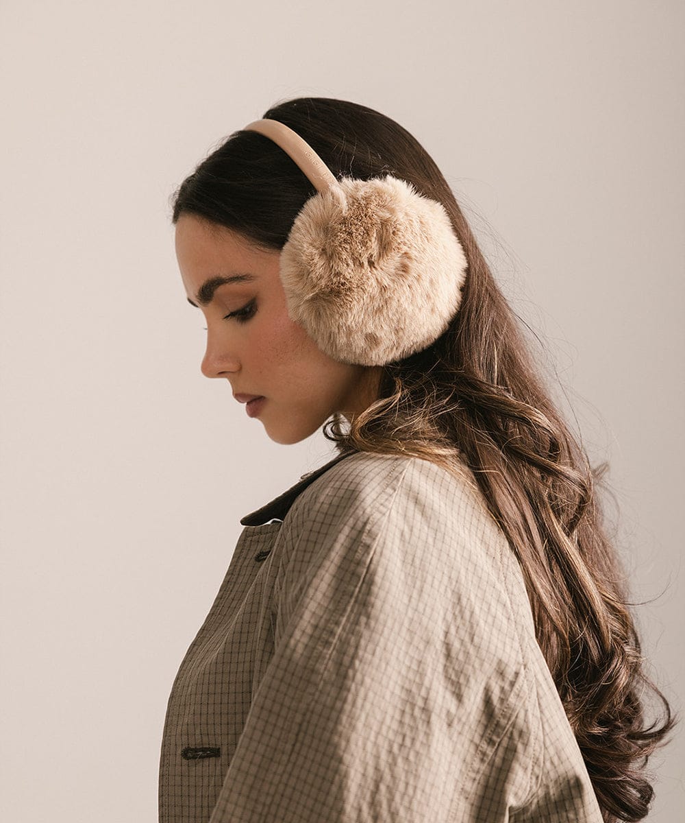 Woman wearing beige fur ear muffs against a neutral background #color_tan