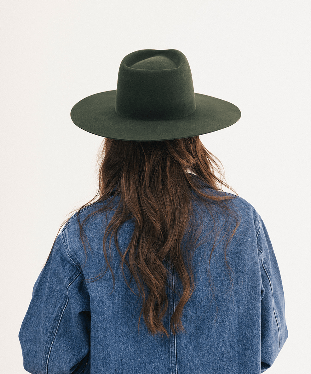 A person viewed from behind wearing a dark olive green wide-brim fedora hat and a blue denim shirt against a plain white background #color_dark green