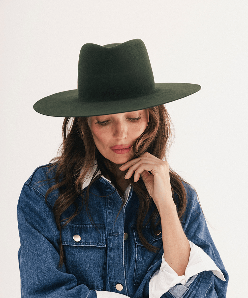 A woman wearing a dark forest green wide-brim fedora hat and a blue denim jacket over a white shirt, posing against a clean off-white background #color_dark green