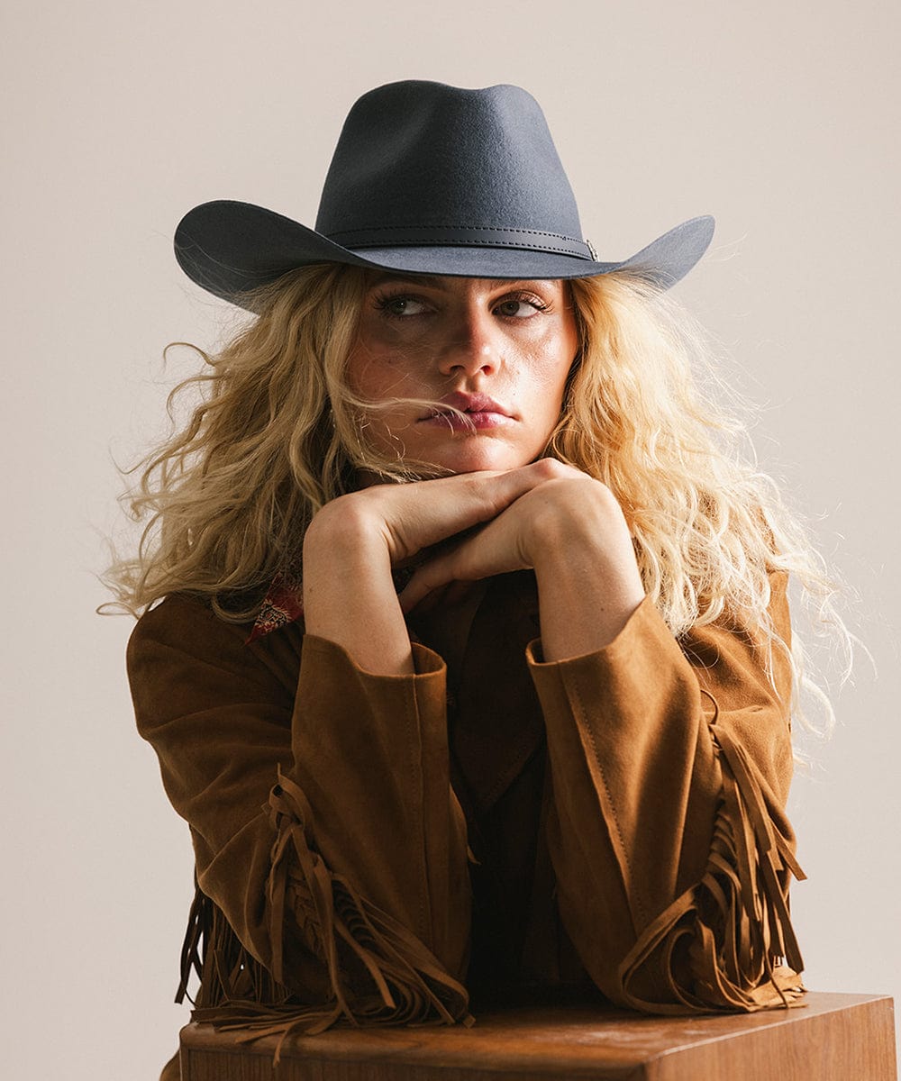 Woman wearing a cowboy hat and brown jacket with fringe, sitting on a wooden block against a plain background #color_vintage blue