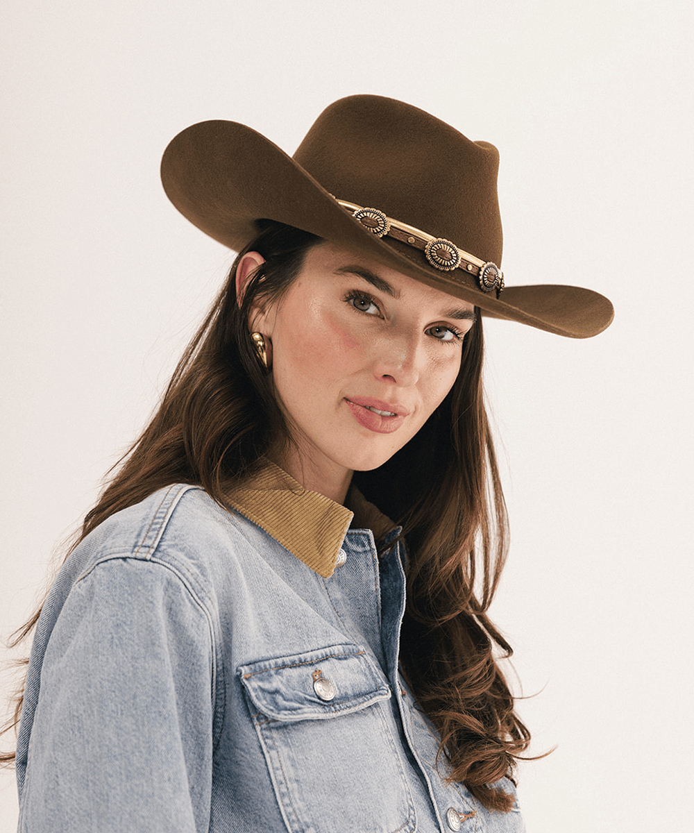 A woman wearing a brown cowboy hat with a decorative metallic band and a light blue denim jacket, photographed against a plain light background #color_coffee