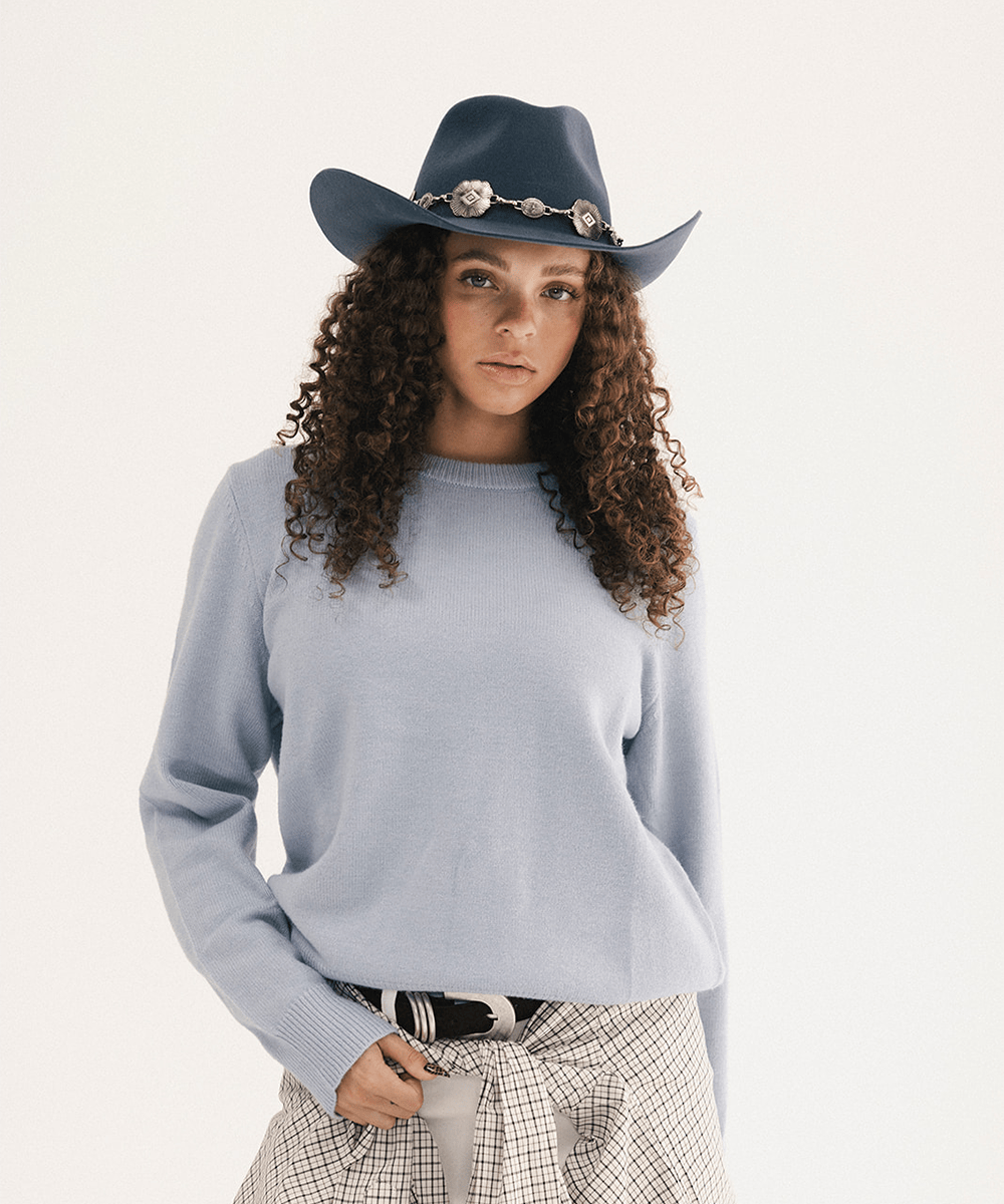 Woman wearing a blue cowboy hat with a concho hat band and a light blue sweatshirt with a plaid shirt tied around the waist #color_vintage blue
