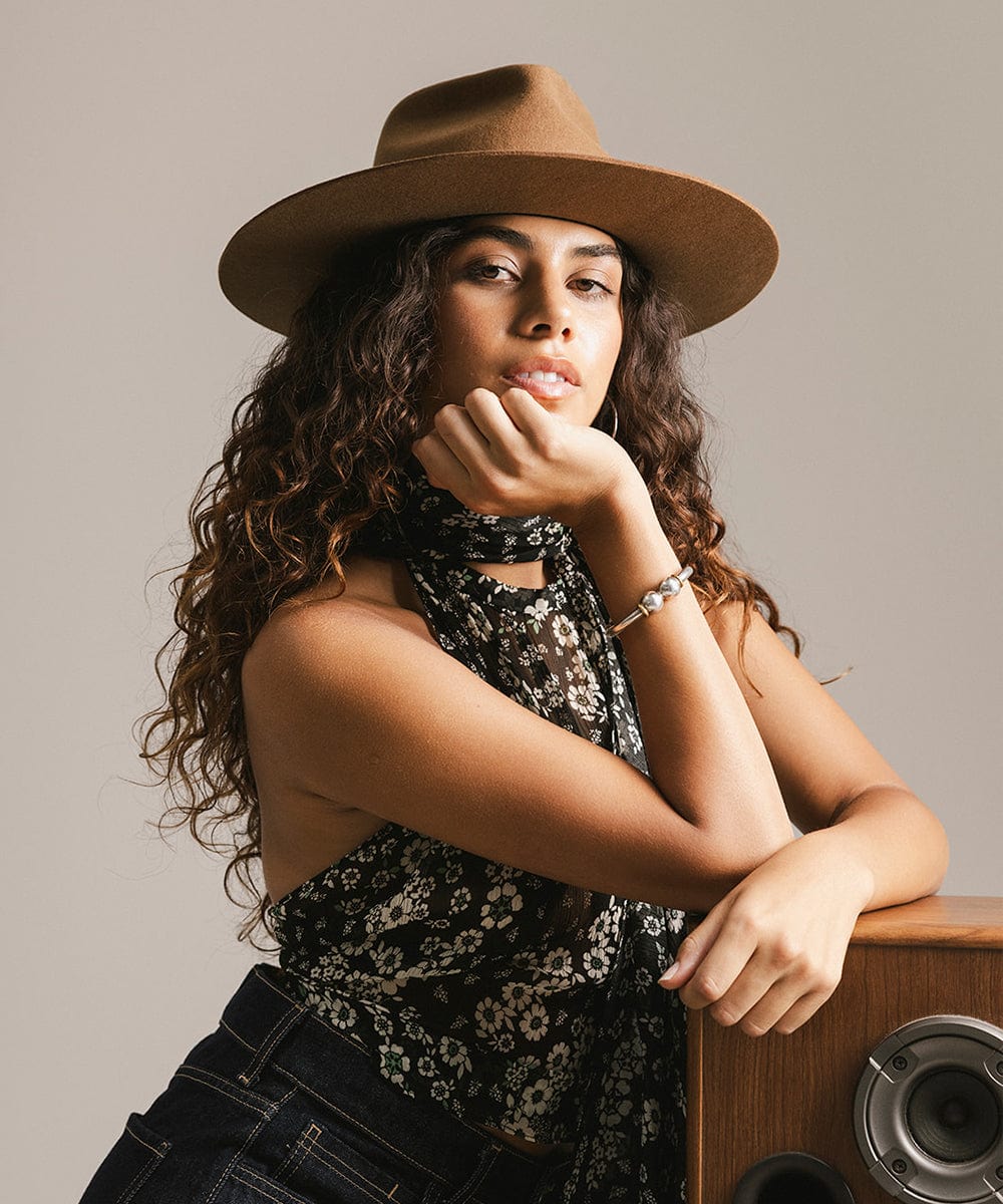 Woman wearing a brown hat and floral scarf, leaning against a wooden surface. #color_caramel