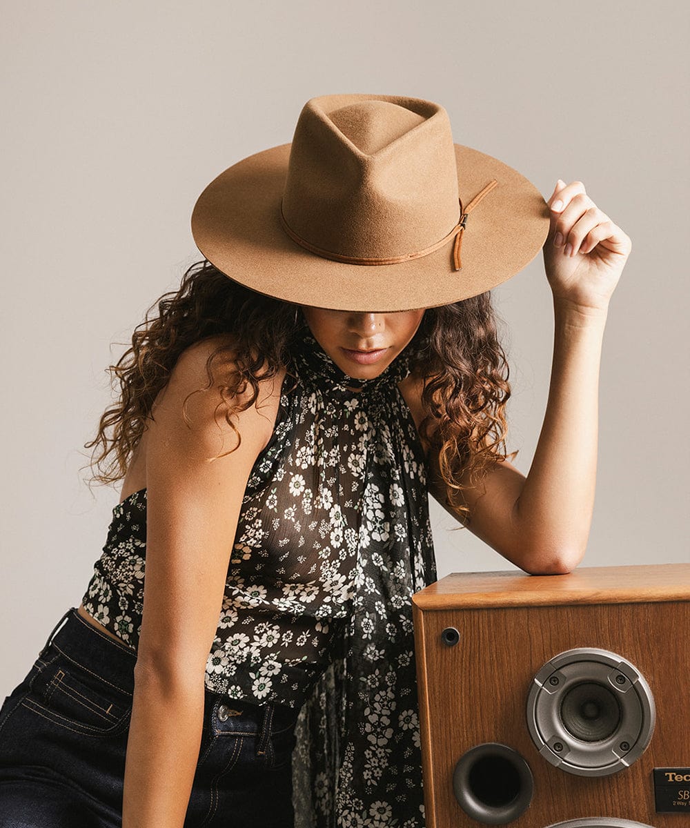 Woman wearing a brown hat and floral scarf, standing next to a wooden speaker. #color_caramel