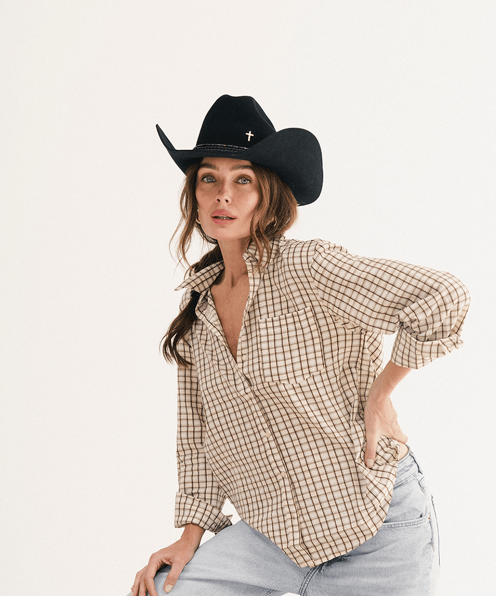 A woman wearing a beige and brown checkered plaid button-down shirt, a black cowboy hat, and light-wash jeans, posing against a solid off-white studio background #color_black