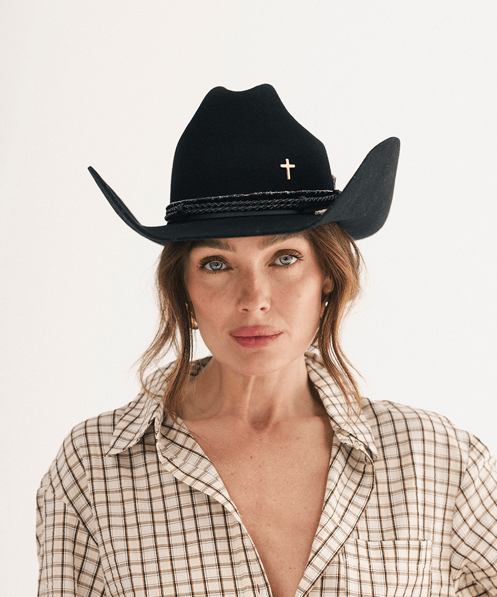A woman wearing a black cowboy hat with a small gold cross and braided band, paired with a beige and brown plaid button-up shirt, against a plain white background #color_black
