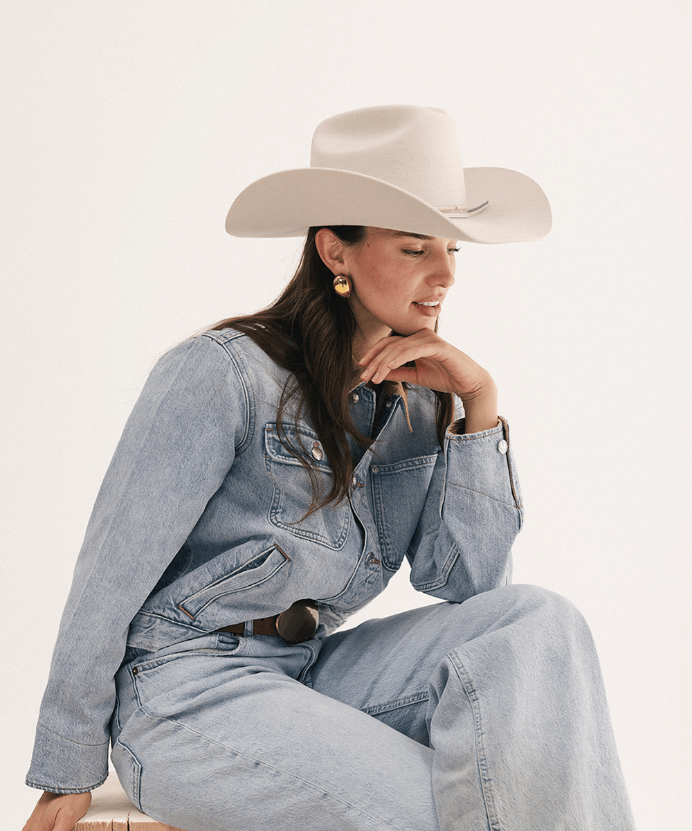 A woman wearing a light beige cowboy hat, a light blue denim jumpsuit, and gold hoop earrings, sitting against a plain off-white studio background #color_off white