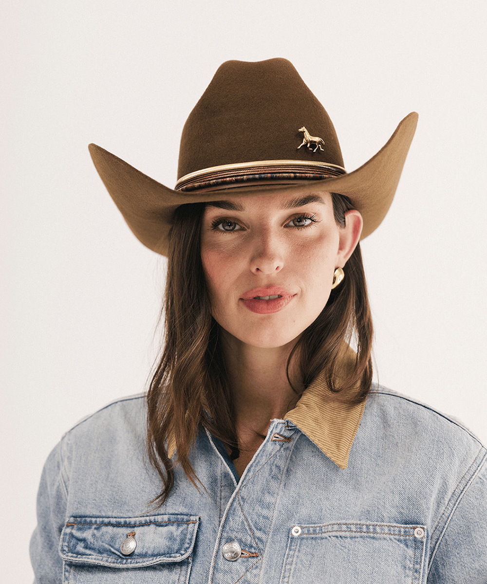 A woman wearing a brown felt cowboy hat with a small gold deer pin on the band, paired with a light blue denim jacket with a tan corduroy collar, against a plain white background #color_coffee
