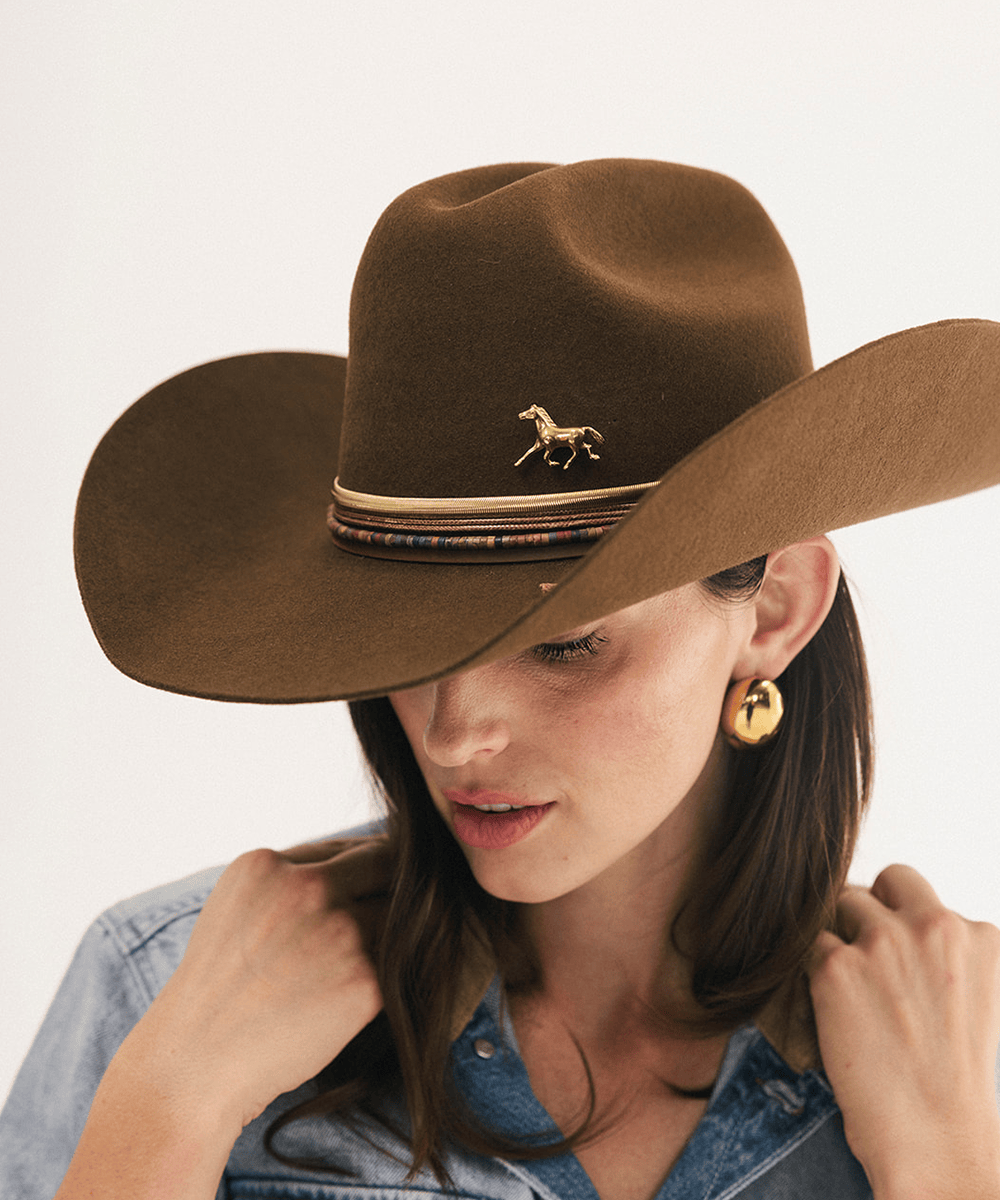 A woman wearing a brown felt cowboy hat with a gold horse emblem and patterned band, paired with a denim shirt against a plain light background #color_coffee