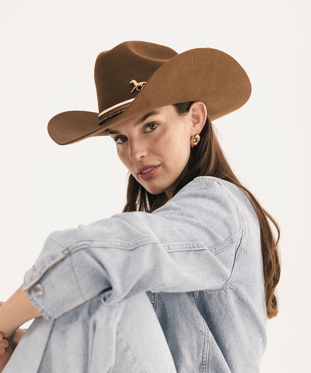 A woman wearing a brown cowboy hat with a small gold horse emblem and a light blue denim shirt, photographed against a clean white studio background #color_coffee