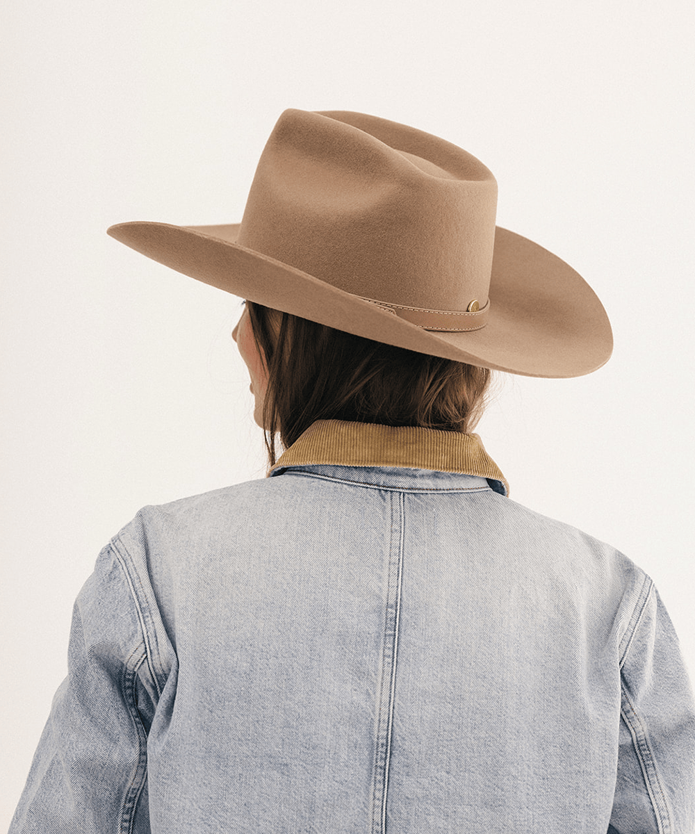 A person shown from behind wearing a tan cowboy hat, a light blue denim jacket with a tan collar, and dark brown hair, photographed against a plain off-white background #color_brown
