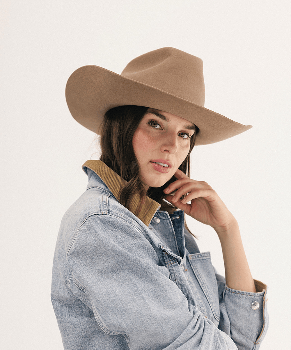 A woman wearing a tan wide-brim felt cowboy hat and a light blue denim jacket with a tan corduroy collar, photographed against a clean off-white studio background #color_brown