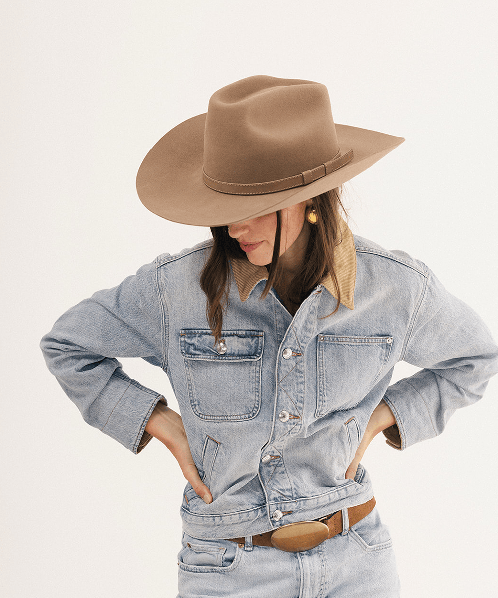 A model wearing a light-wash denim shirt-jacket with a tan collar, styled with a brown wide-brim hat and a brown belt, against a plain off-white background #color_brown