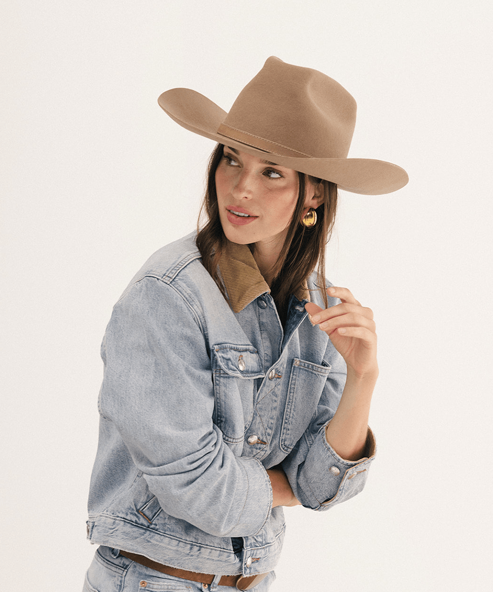 A woman wearing a tan felt cowboy hat, a light-wash denim jacket with a tan collar, and gold hoop earrings, photographed against a plain off-white studio background #color_brown