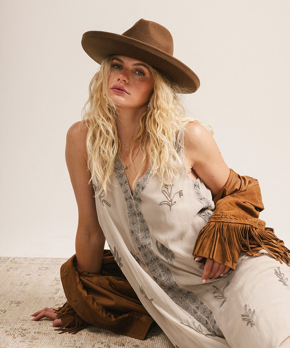 Woman wearing a brown hat and a white dress with floral patterns, sitting on a textured surface. #color_chocolate