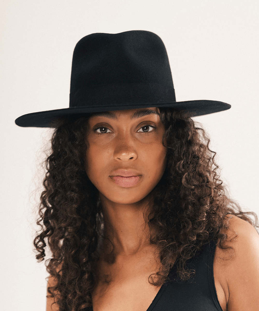 A woman with long, curly brown hair wearing a black wide-brim fedora hat and a black sleeveless top, photographed against a plain light background #color_black