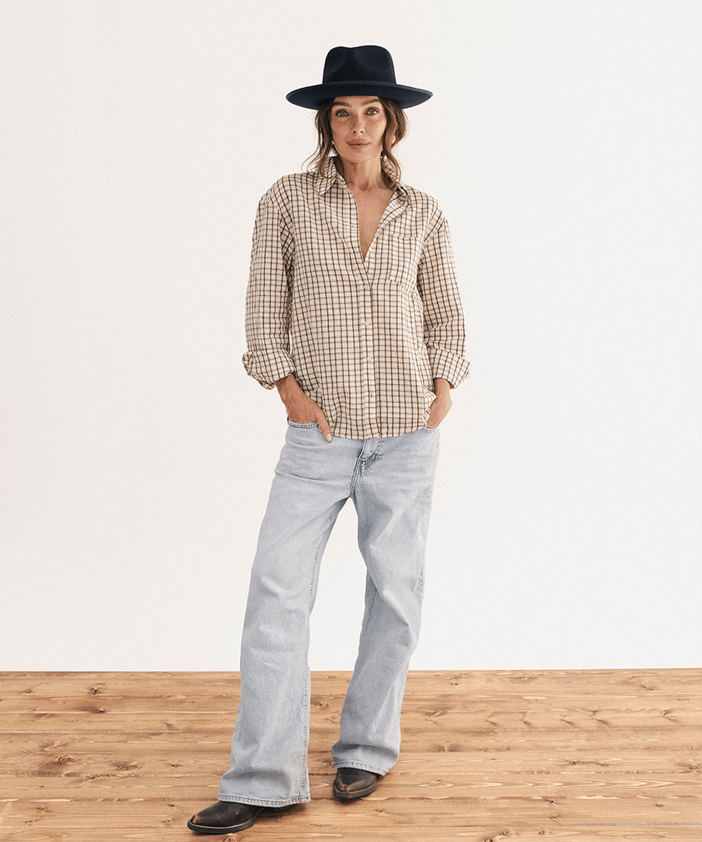 A woman stands in a studio wearing a beige and brown gingham button-up shirt, light-wash wide-leg jeans, a black wide-brim hat, and black shoes, with her hands in her jeans pockets #color_navy