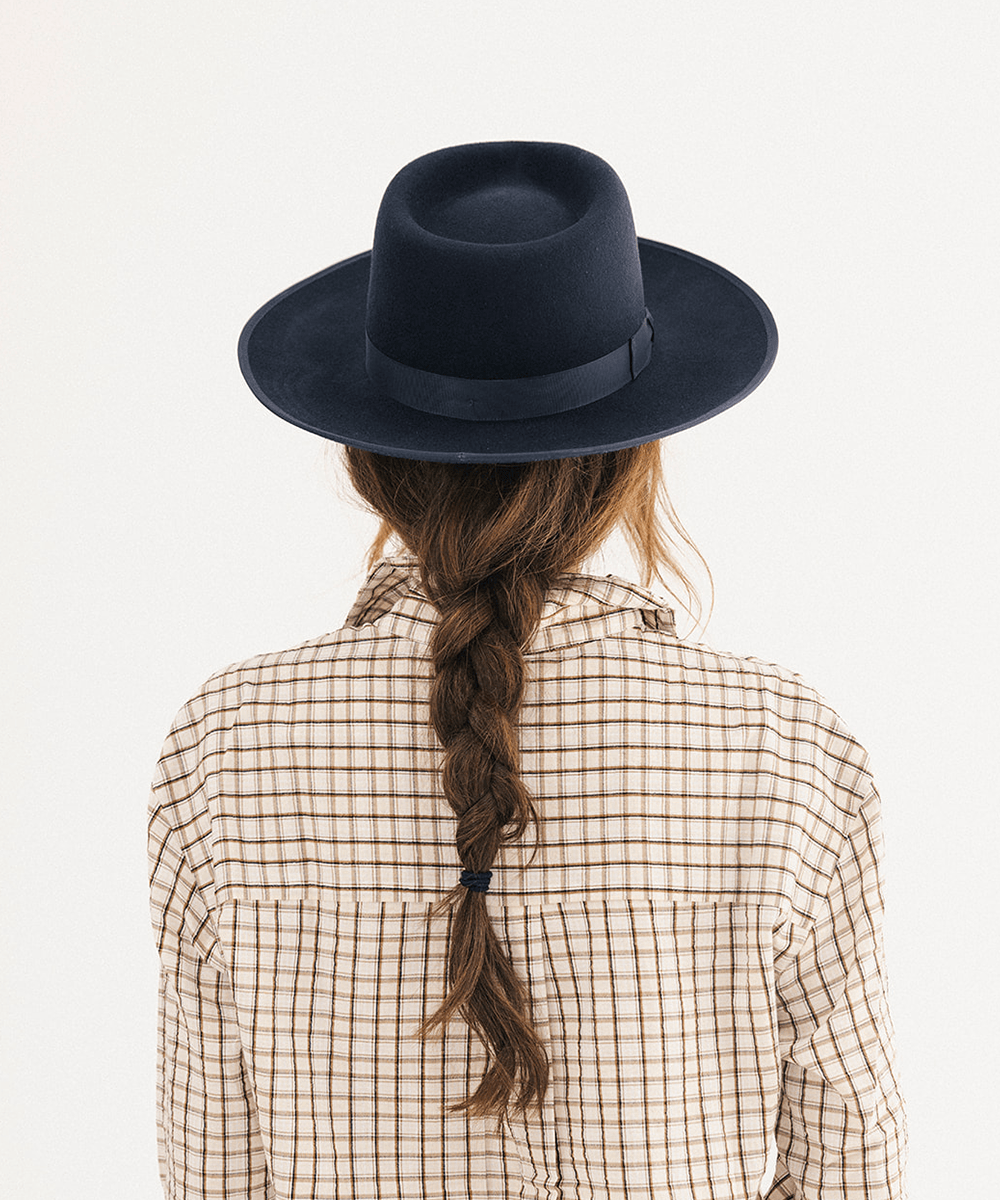 A person viewed from behind wearing a dark navy blue felt fedora hat with a matching band, a beige and brown checkered shirt, and a long brown braid against a plain white background #color_navy