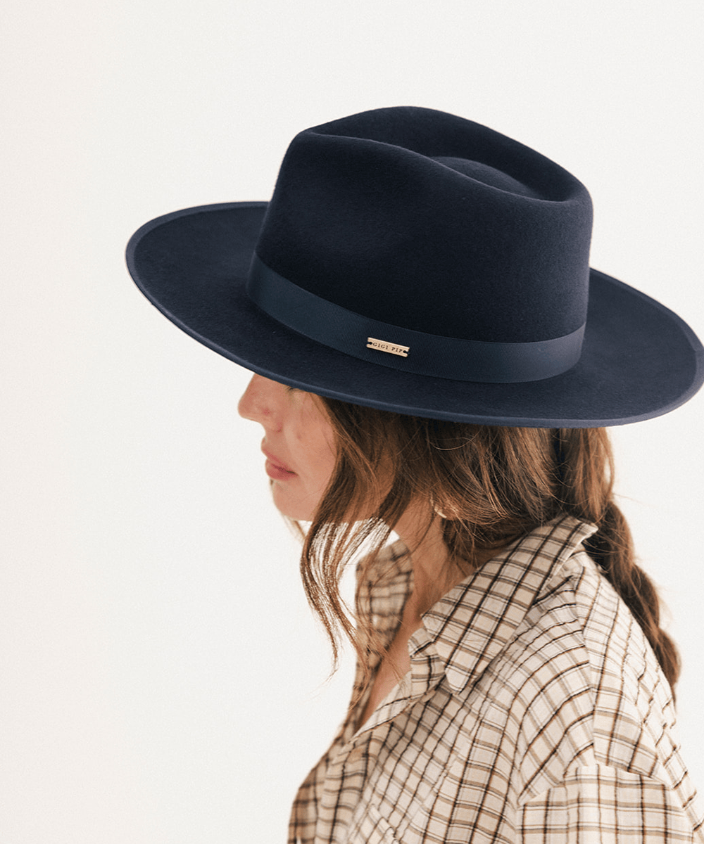 A woman in profile wearing a dark navy blue felt fedora hat with a matching navy band and a small gold logo plaque, paired with a beige and brown plaid shirt against a plain white background #color_navy