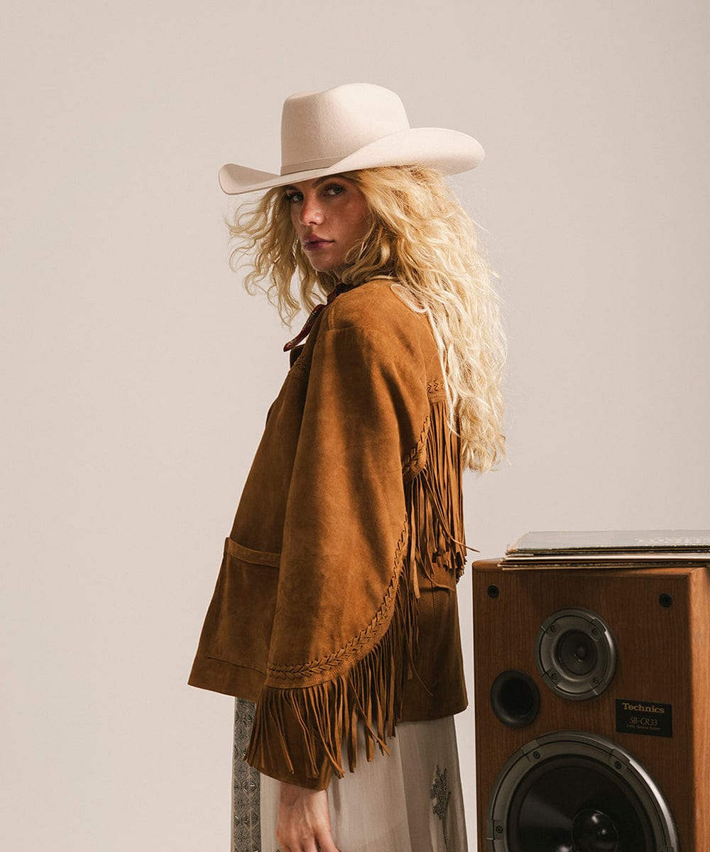 Woman wearing a brown fringed jacket and cowboy hat standing next to a wooden speaker. #color_cream