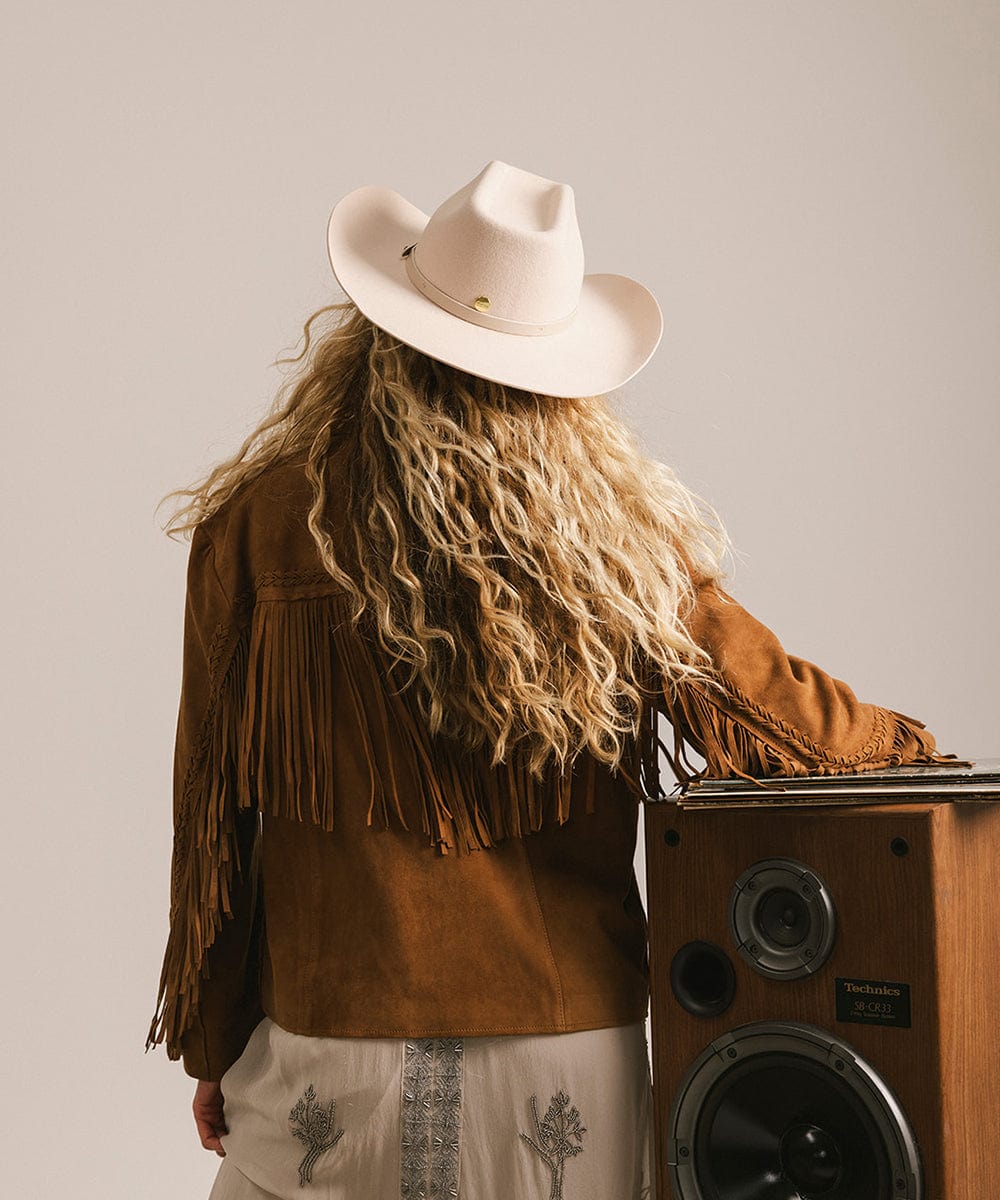 Person wearing a cowboy hat and brown fringed jacket standing next to a wooden speaker. #color_cream