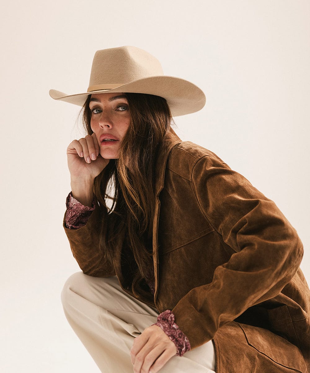 Woman wearing a brown suede jacket and beige cowboy hat against a plain background #color_tan