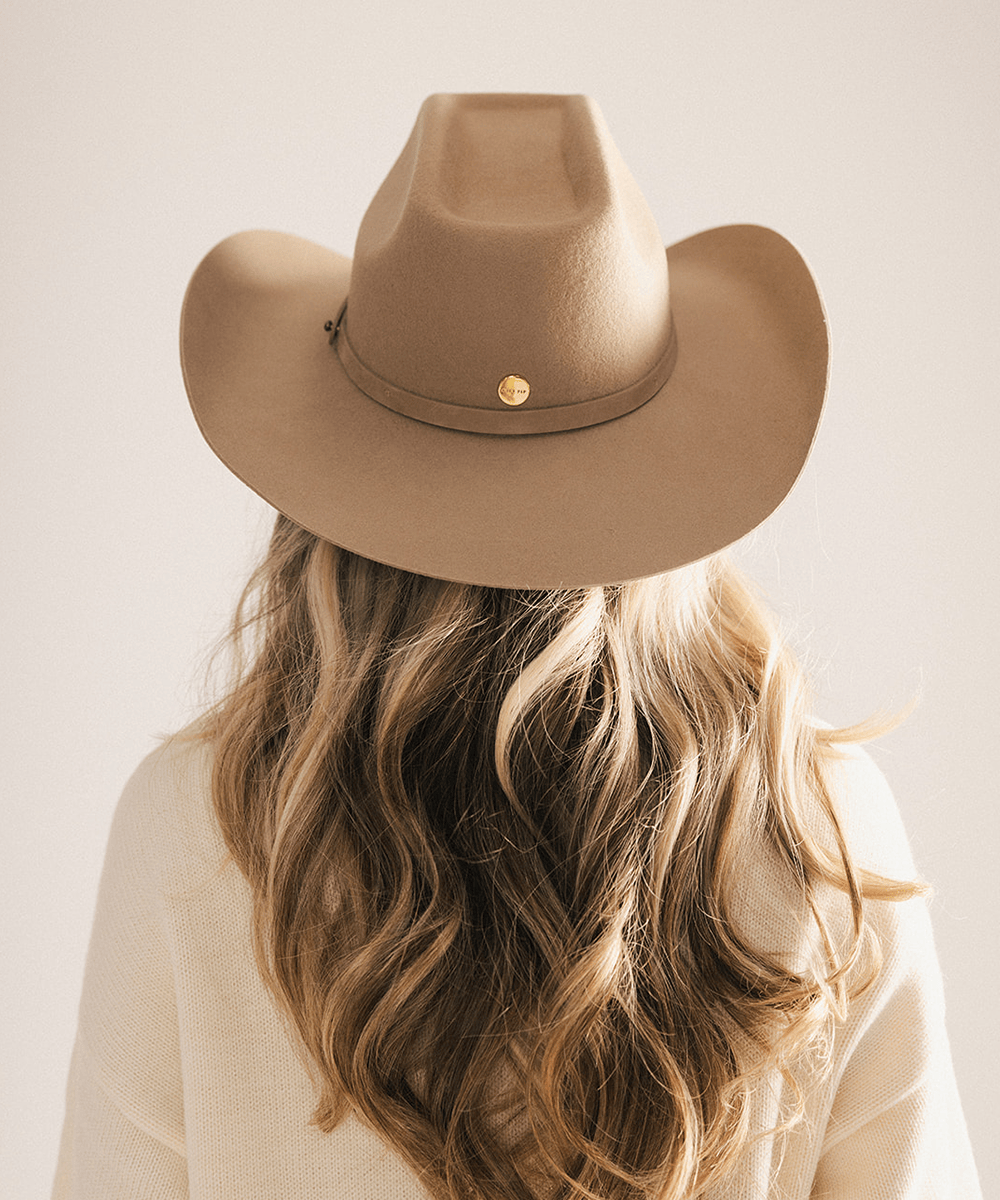 A woman viewed from behind wearing a tan cowboy hat with a small gold button and a cream knit top, set against a plain light beige background #color_tan