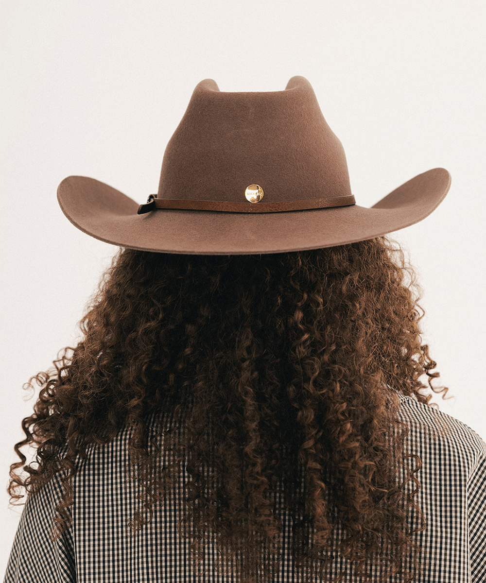 Woman wearing a chocolate colored cowboy hat with an upturned brim and an attached leather band with a star pin, displayed against a white background. #color_chocolate