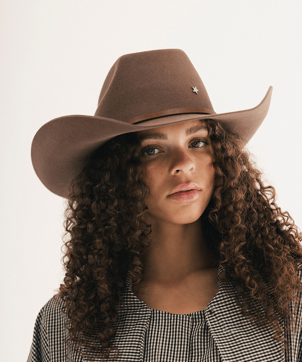 Woman wearing a chocolate colored cowboy hat with an upturned brim and an attached leather band with a star pin, displayed against a white background. #color_chocolate