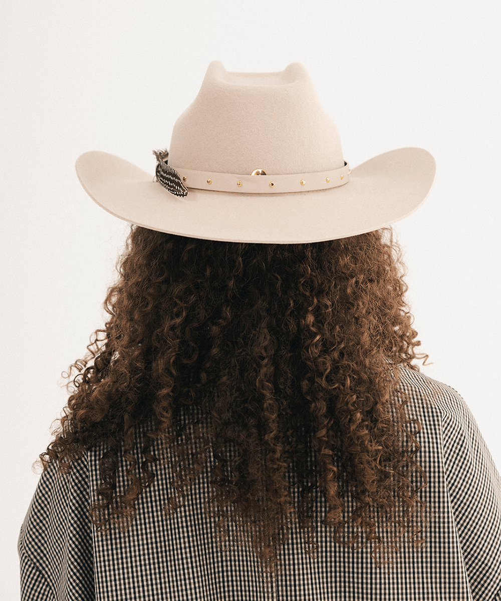Woman wearing a cream cowboy hat with an upturned brim and an attached cream leather band, feather, and a plaid shirt on a white background #color_cream
