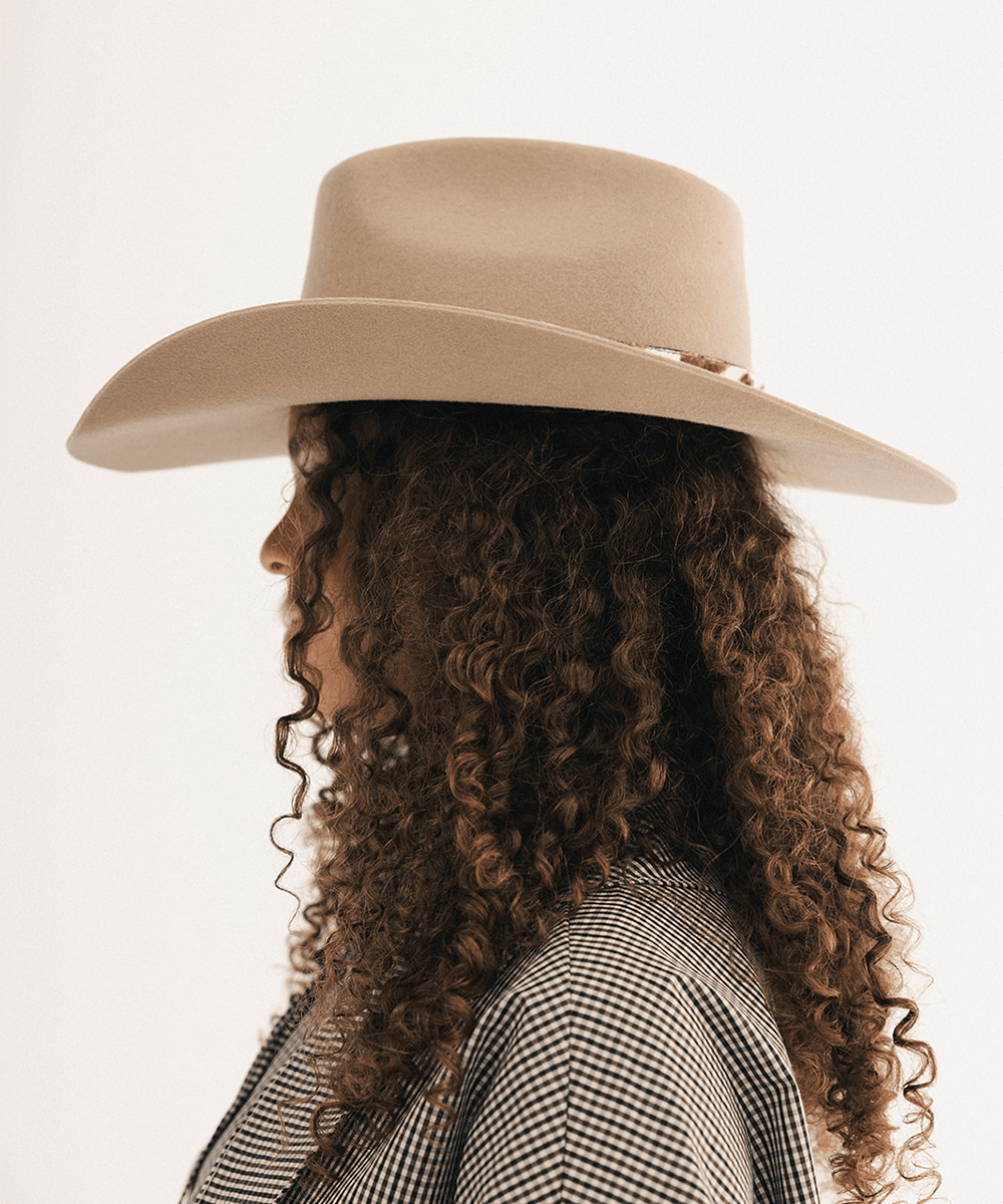 Woman wearing a tan cowboy hat with an upturned brim and an attached cow leather band, displayed against a white background #color_tan