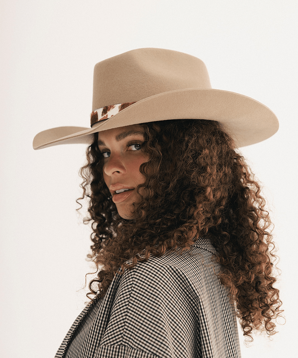 Woman wearing a tan cowboy hat with an upturned brim and an attached cow leather band, displayed against a white background #color_tan