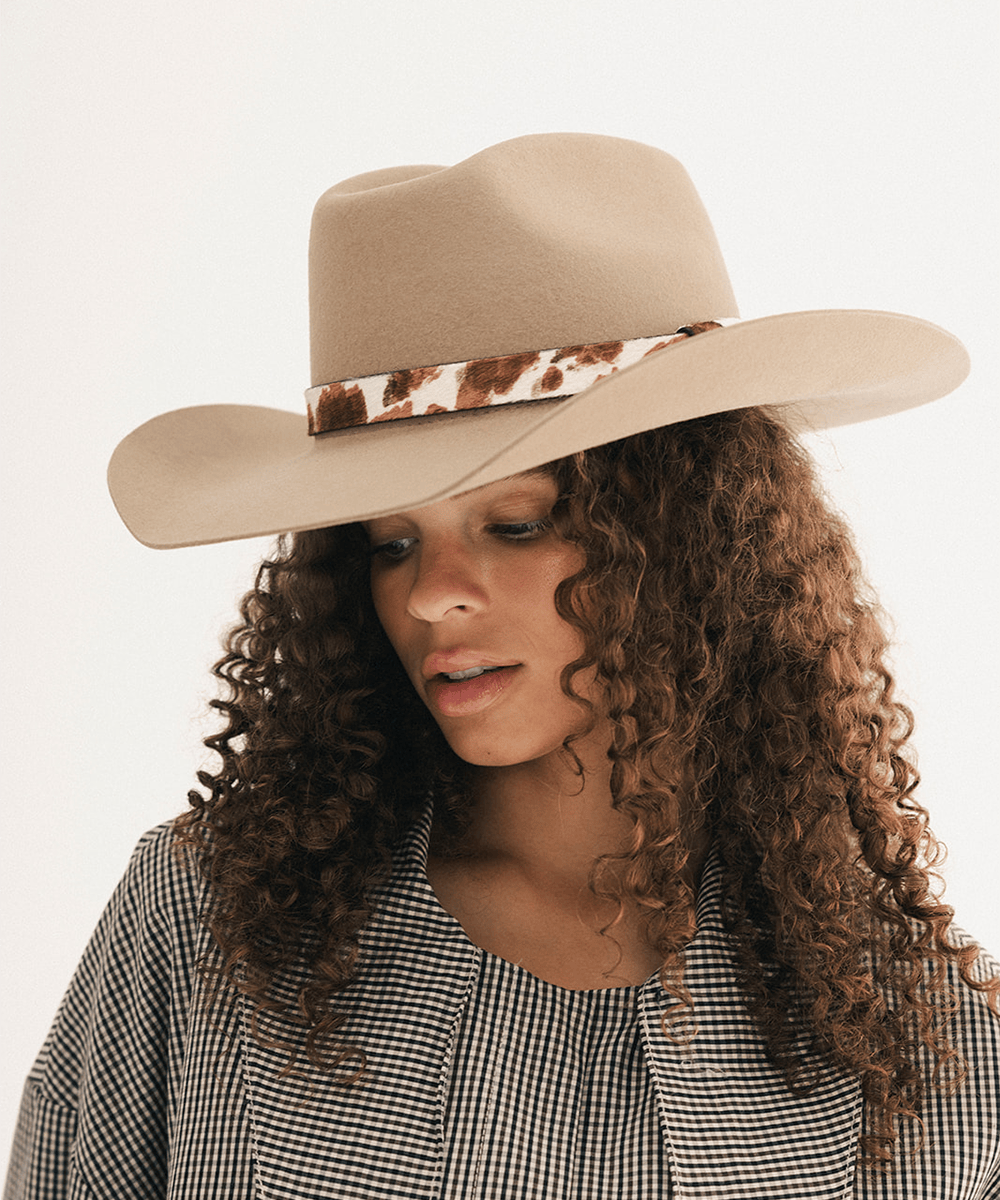 Woman wearing a tan cowboy hat with an upturned brim and an attached cow leather band, displayed against a white background #color_tan