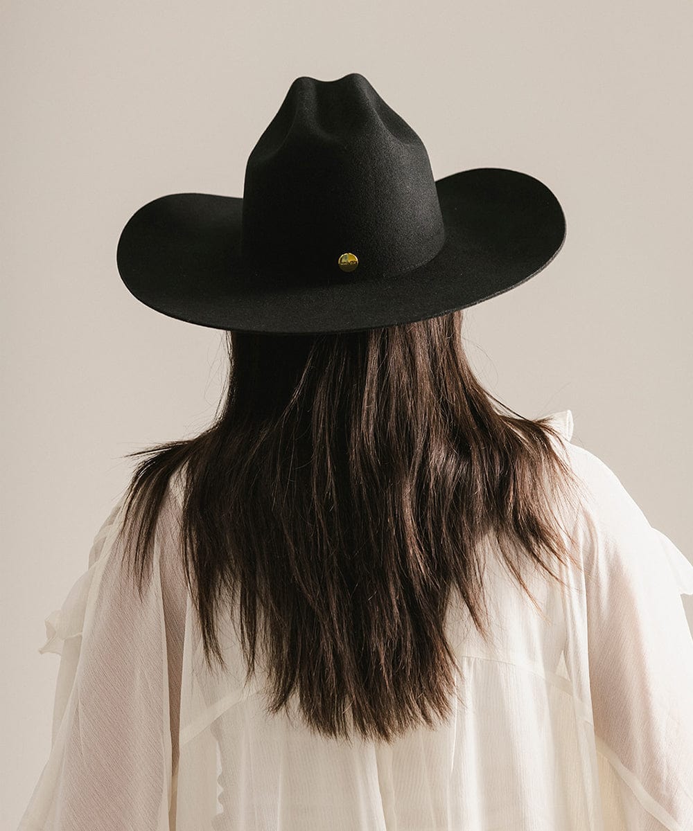 Woman wearing a black cowboy hat with a gold button on a plain background #color_black