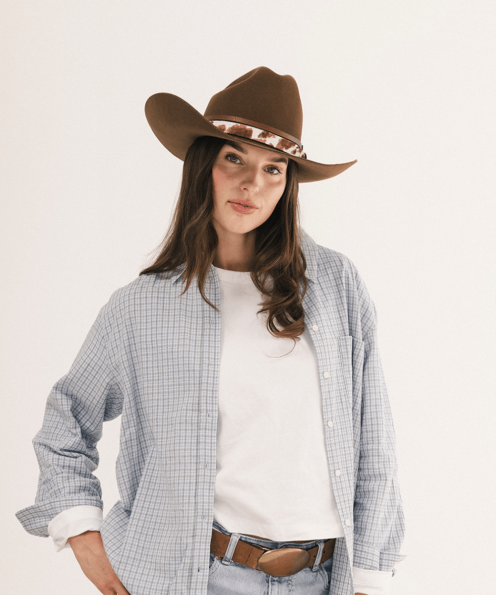 A woman wearing a brown cowboy hat with a patterned band, a light blue and white checkered button-up shirt over a white t-shirt, and jeans with a brown belt, photographed against a plain white studio background #color_coffee